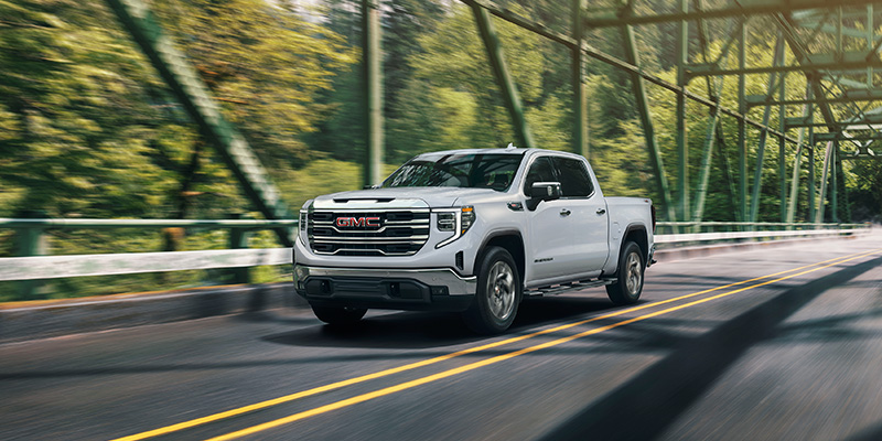 A white 2026 GMC Sierra 1500 drives across a green tree-lined bridge in the countryside.